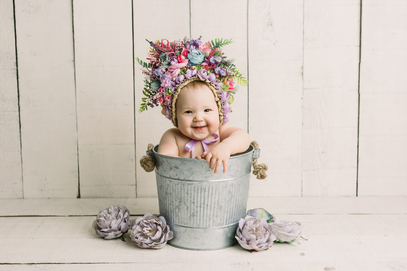 Laughing baby in colorful floral bonnet sitting in metal bucket, Boise baby photographer